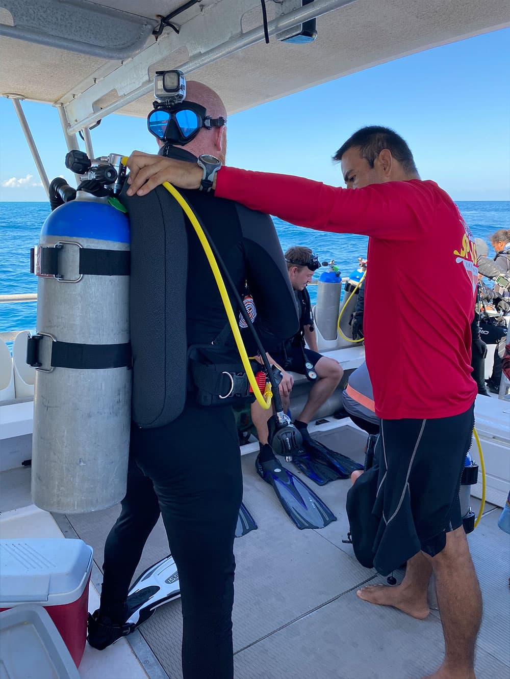 Two divers prepare for scuba diving on a boat, one assisting the other with equipment.