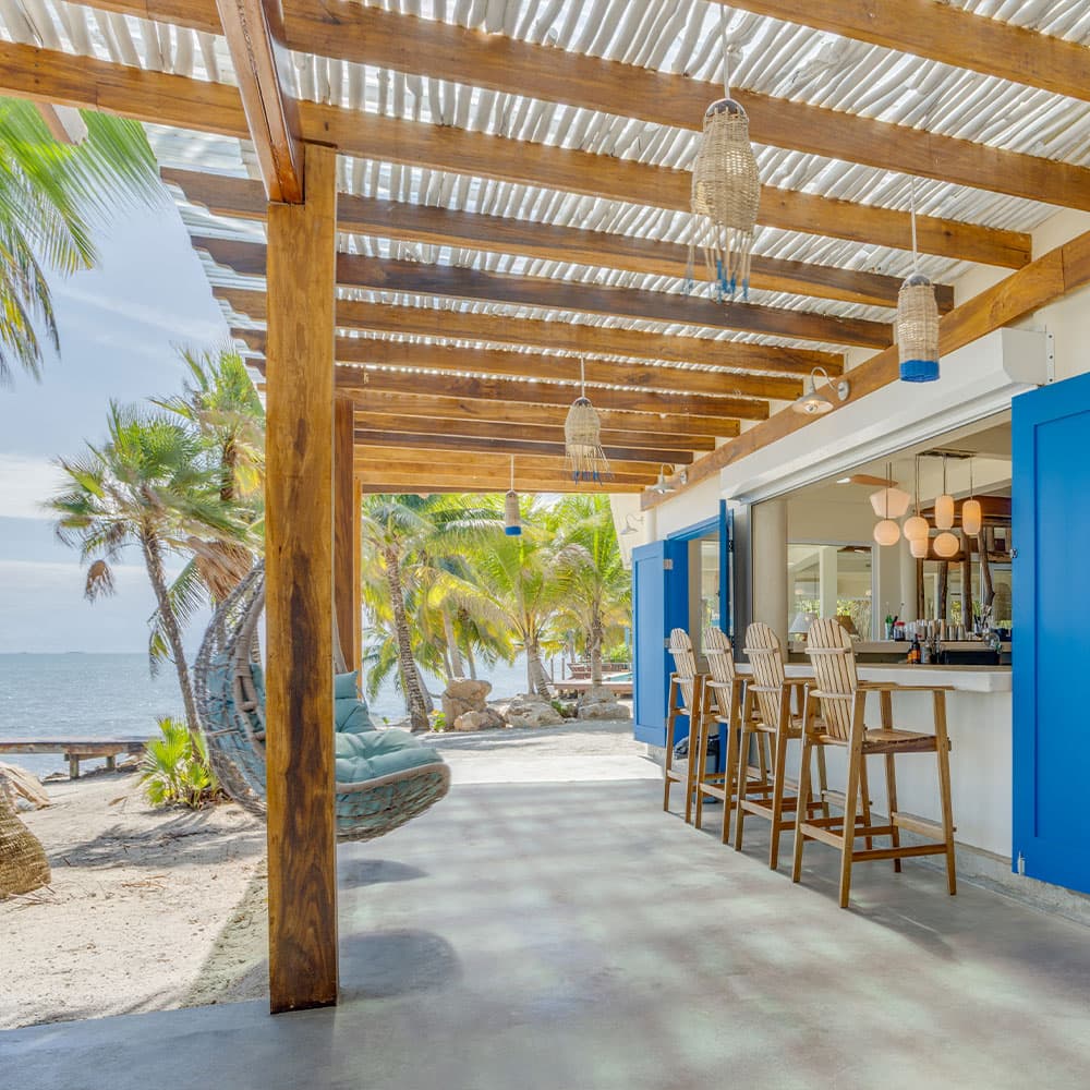 A beachfront bar area with wooden posts, seating, and tropical plants under a roof with slatted sunlight.