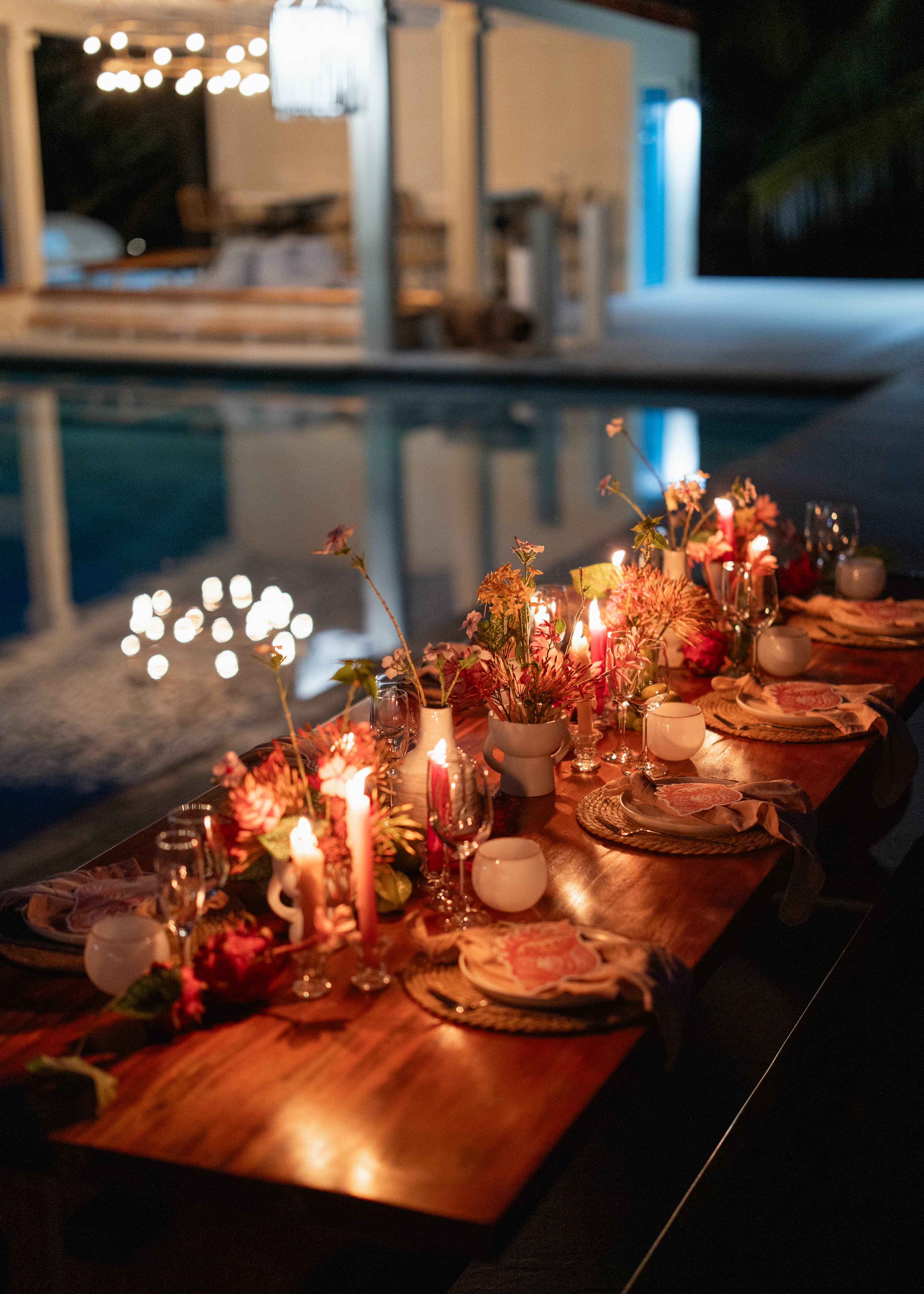 A beautifully decorated outdoor dining table lit by candles beside a serene pool at night.