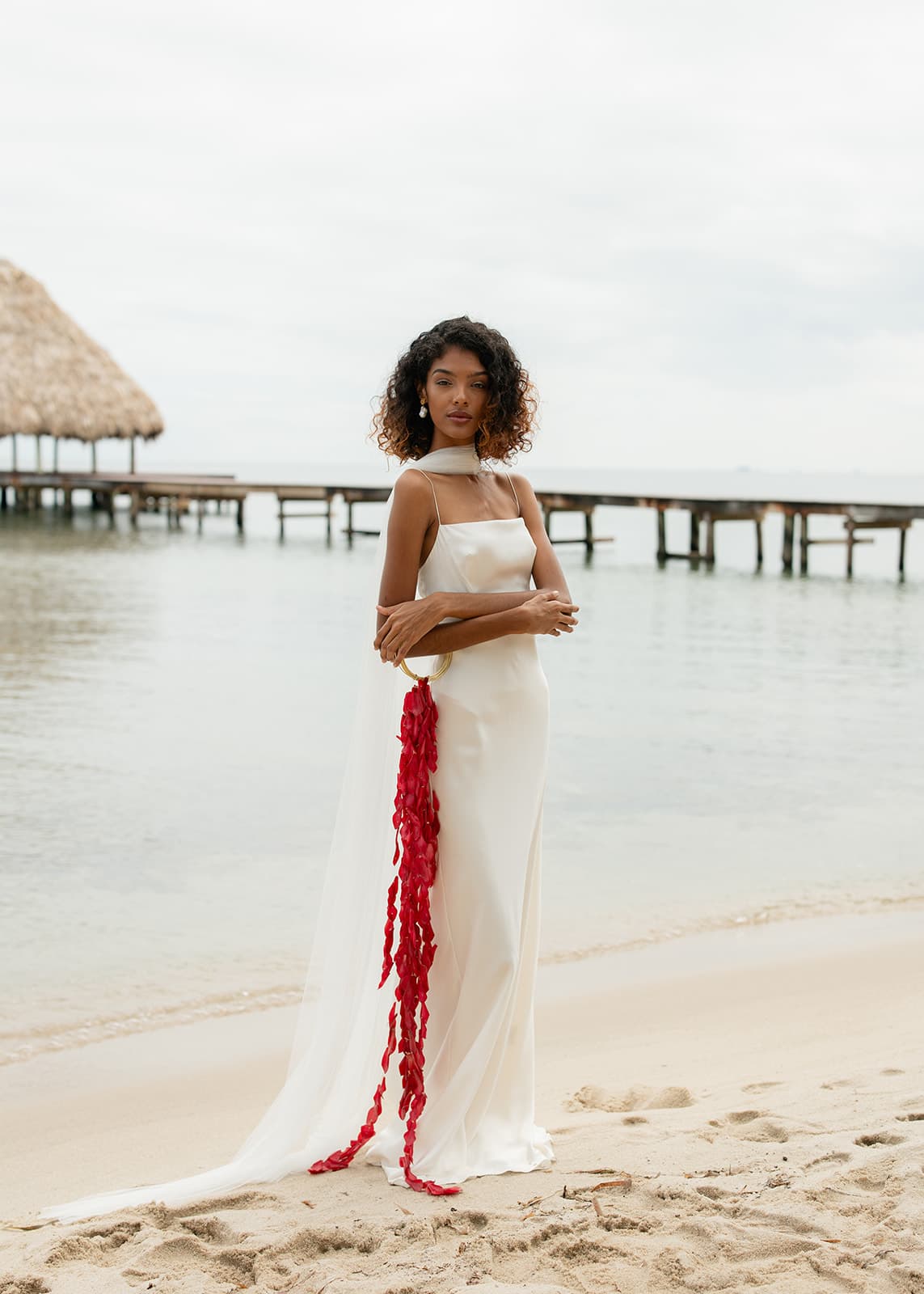 A woman in a white gown with a red detail stands on a beach near an overwater pier.
