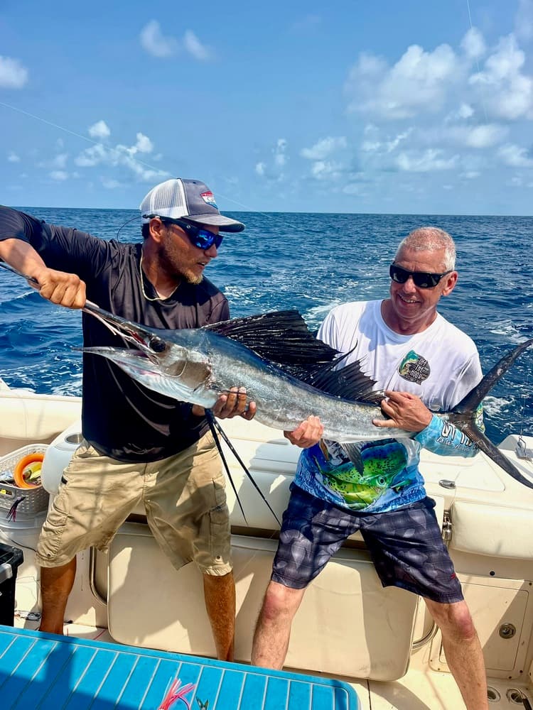 Two men on a boat pose with a large fish against a backdrop of blue ocean and sky.
