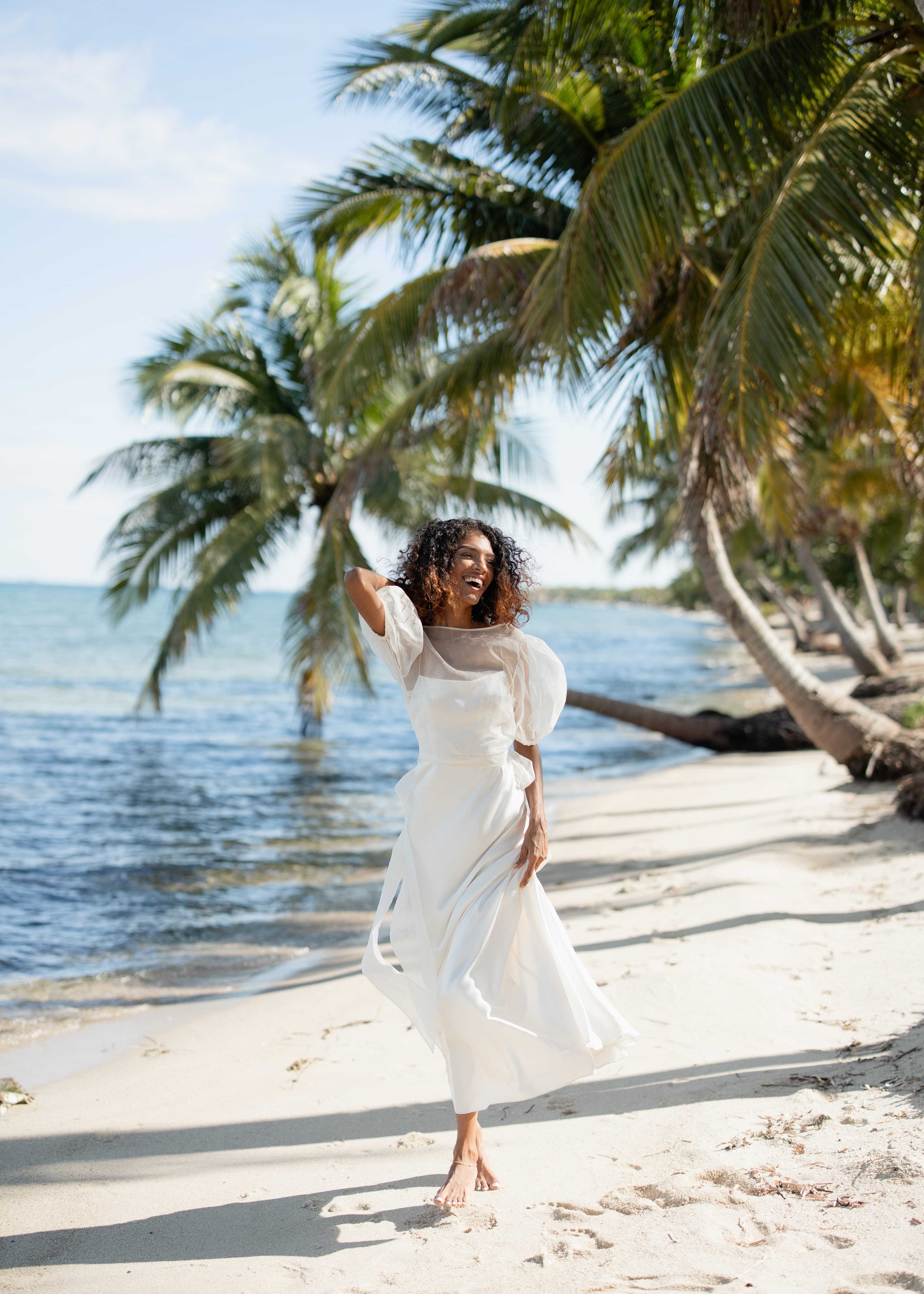 A woman in a white dress joyfully poses on a sandy beach surrounded by palm trees.