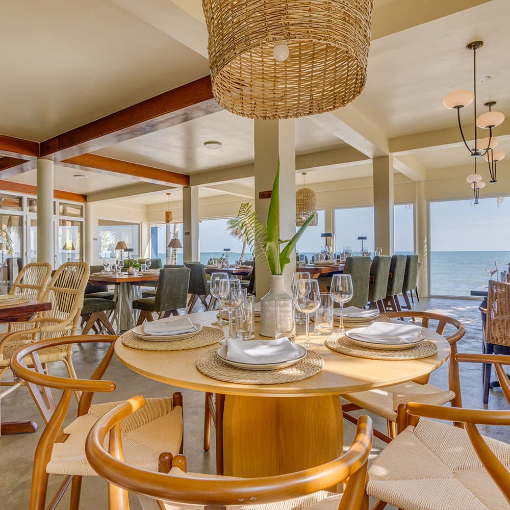 A bright, elegant restaurant with wooden decor, a round table set for dining, and large windows overlooking the sea.