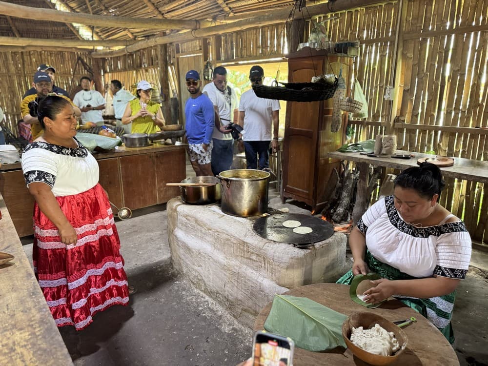 A woman in traditional dress prepares food in a rustic kitchen while others look on.