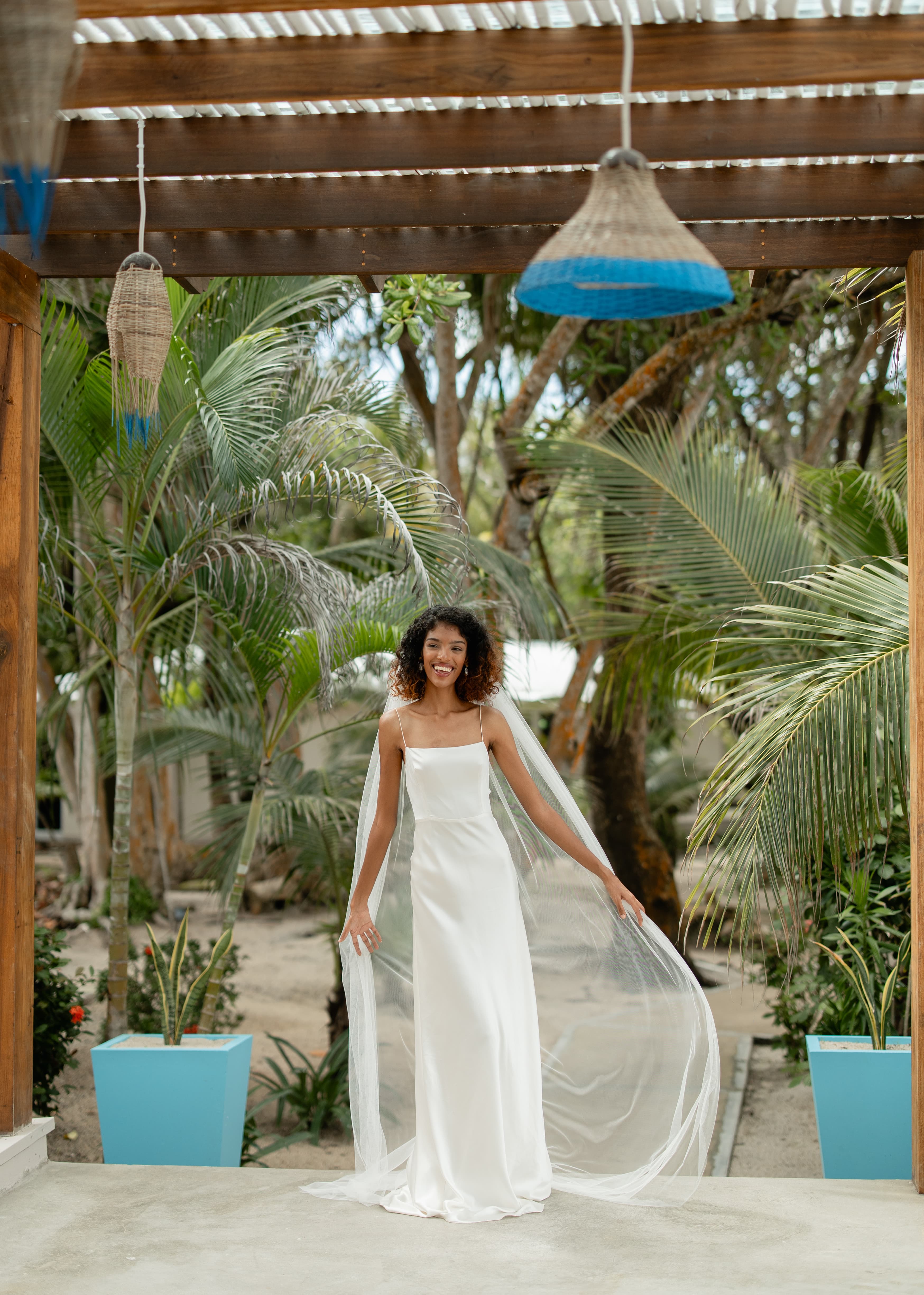 A woman in a flowing white dress stands gracefully with a veil in a tropical setting.