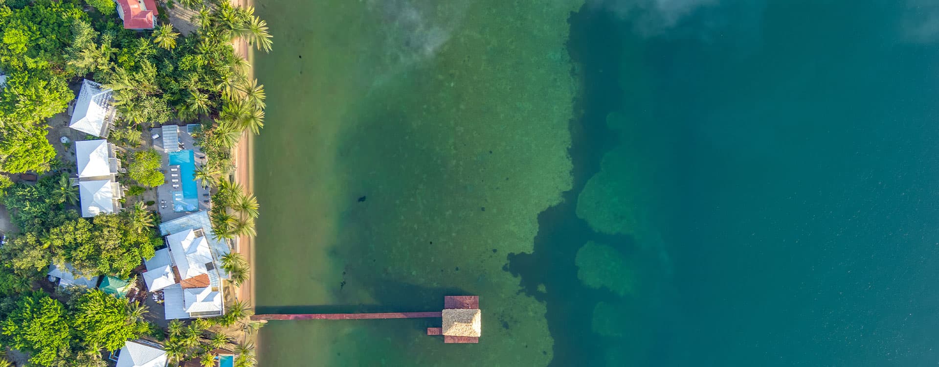 Aerial view of a resort area with bungalows by a clear blue lagoon and a wooden dock.