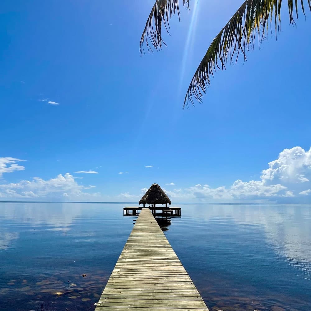 A wooden dock leads to a thatched-roof gazebo over calm water under a bright blue sky.