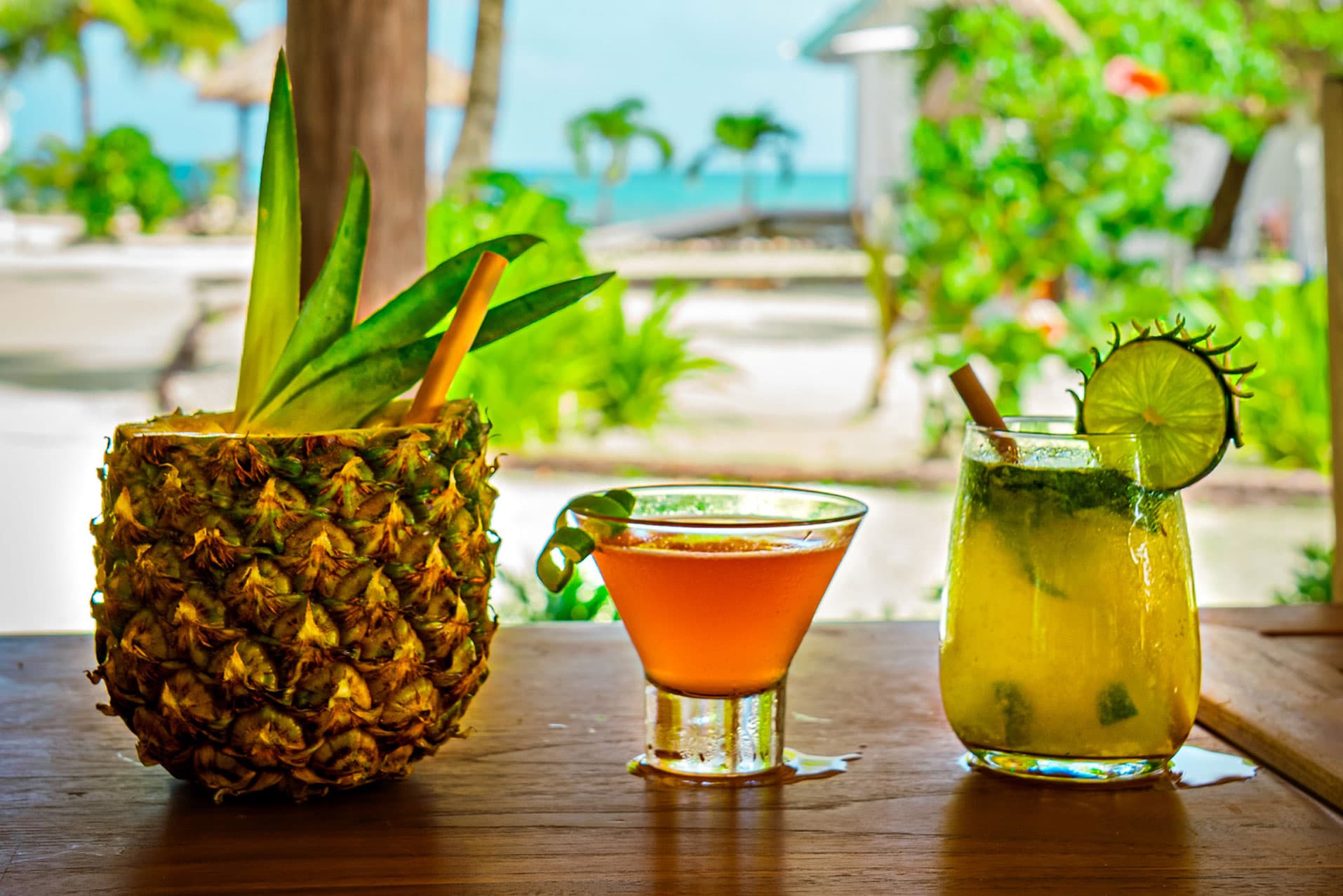Three colorful cocktails sit on a wooden table with a tropical background.