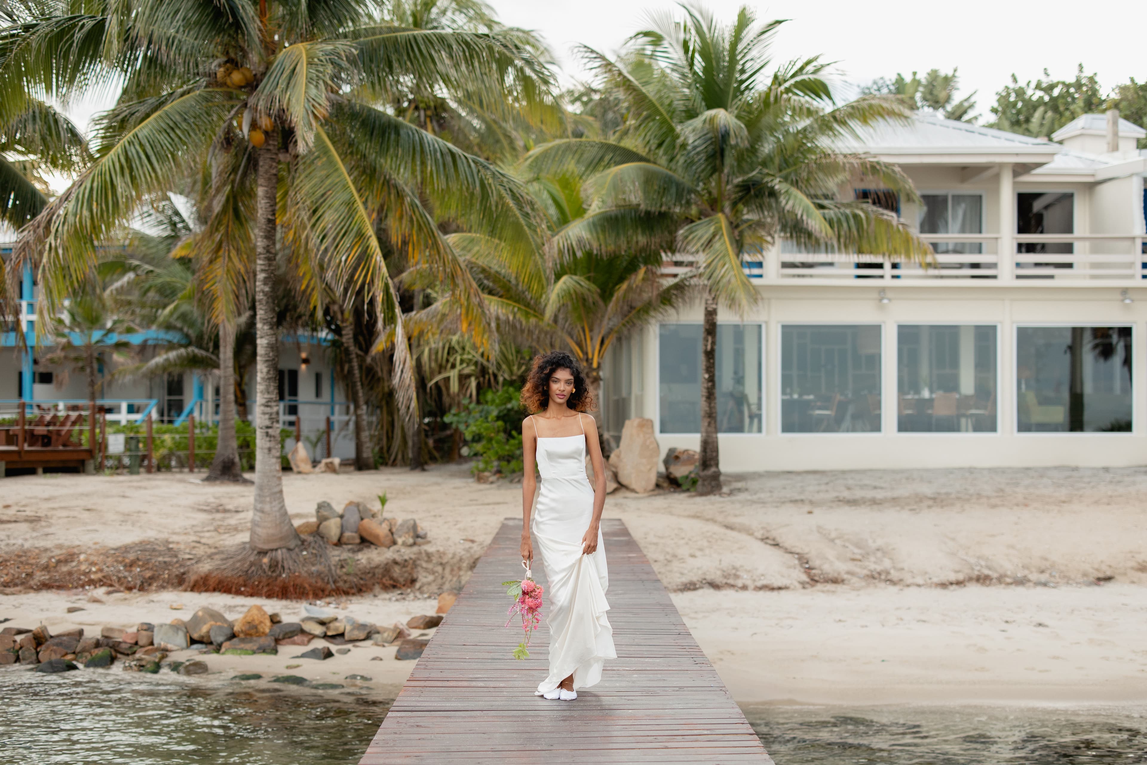 A woman in a white dress walks along a wooden path near the beach, surrounded by palm trees and a house in the background.