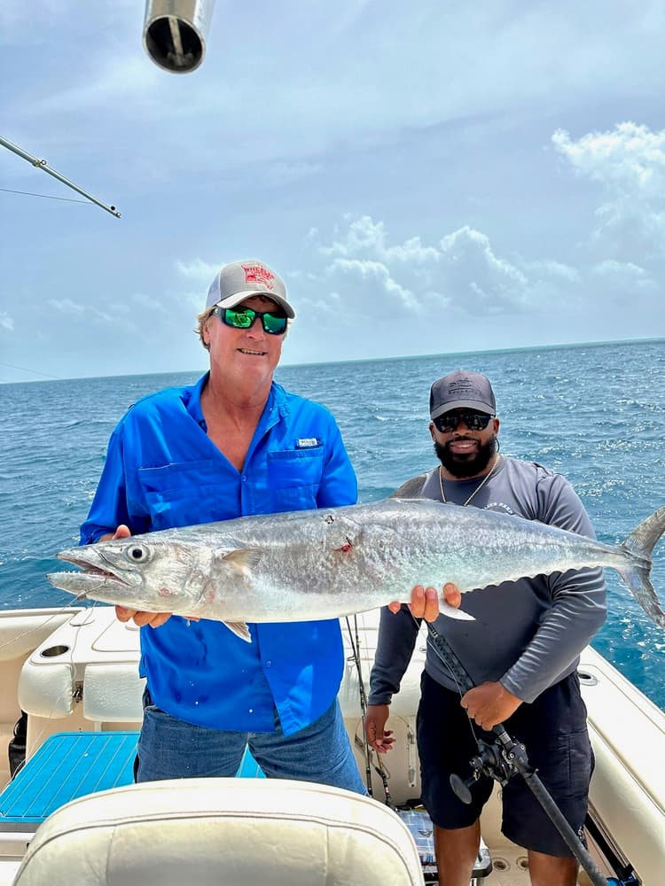 Two men on a boat proudly display a large fish against a backdrop of ocean and cloudy sky.