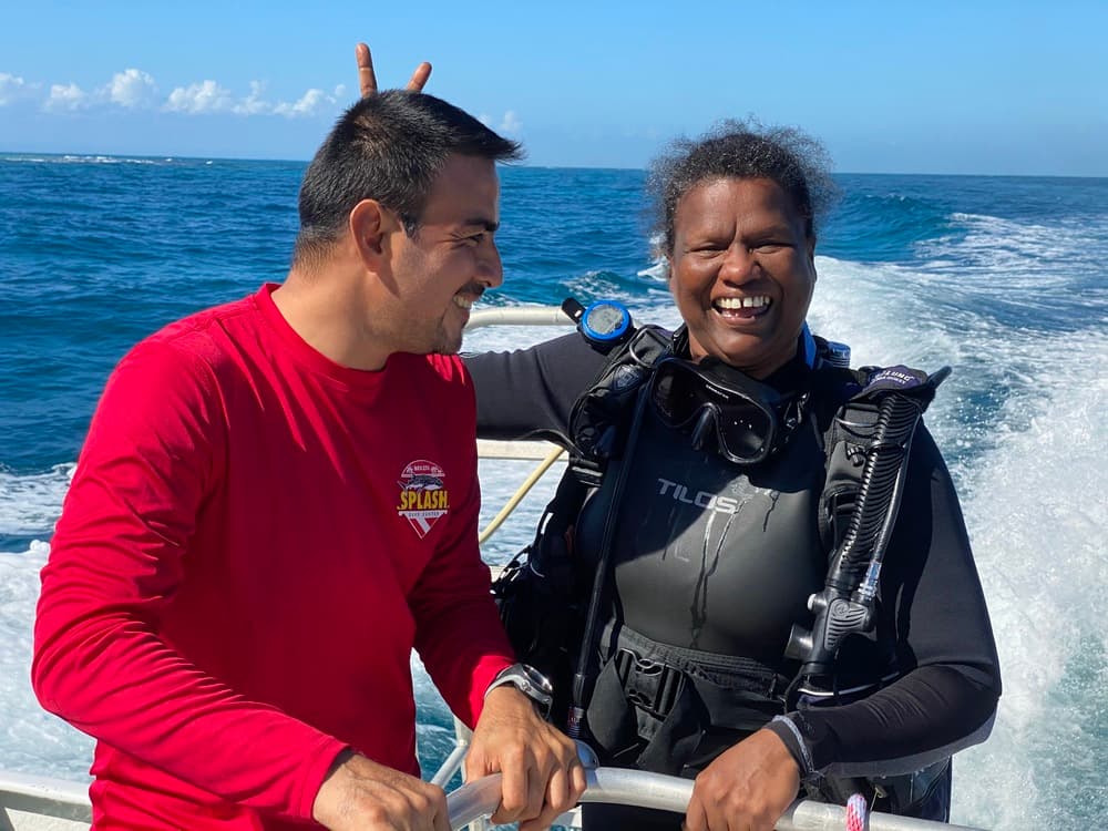 A man playfully makes a peace sign behind a smiling woman in scuba gear, with a clear blue ocean in the background.