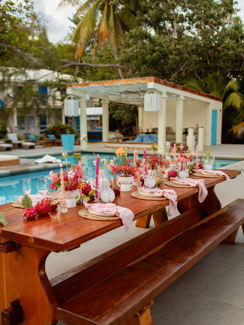 A beautifully arranged outdoor dining table adorned with vibrant flowers and candles, set beside a sparkling pool.