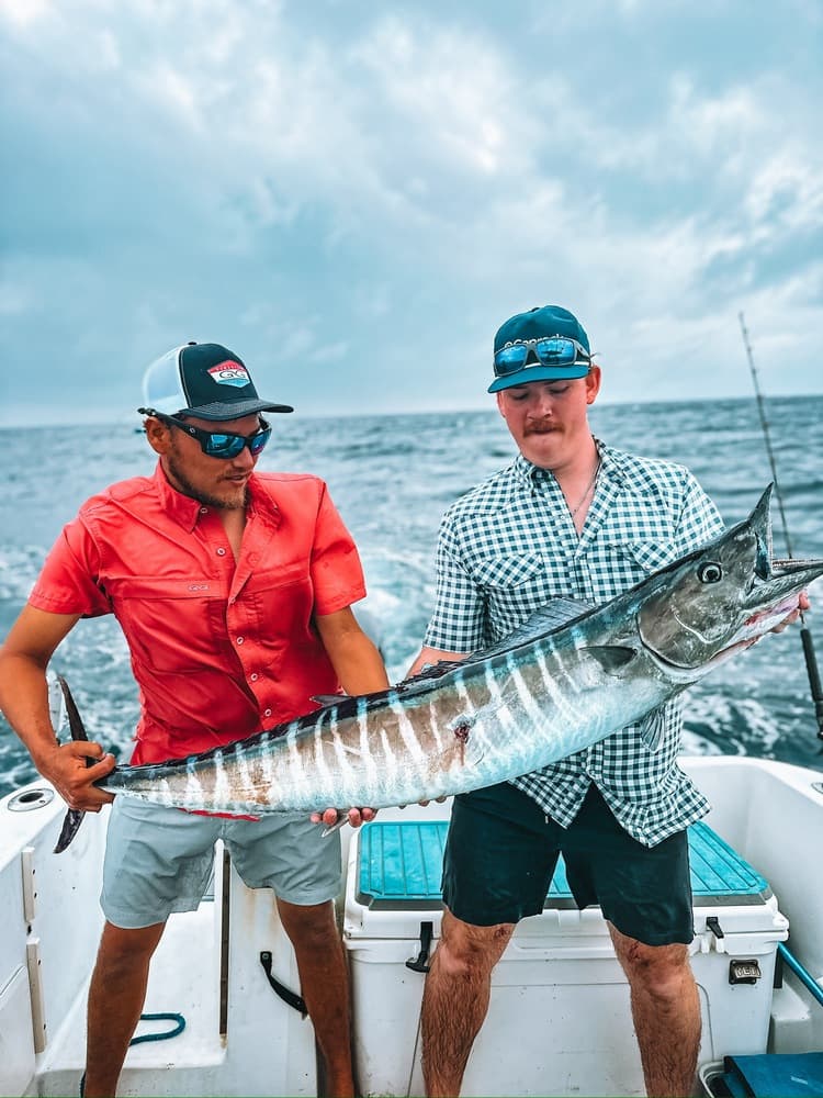 Two men holding a large fish on a boat with a cloudy sky in the background.