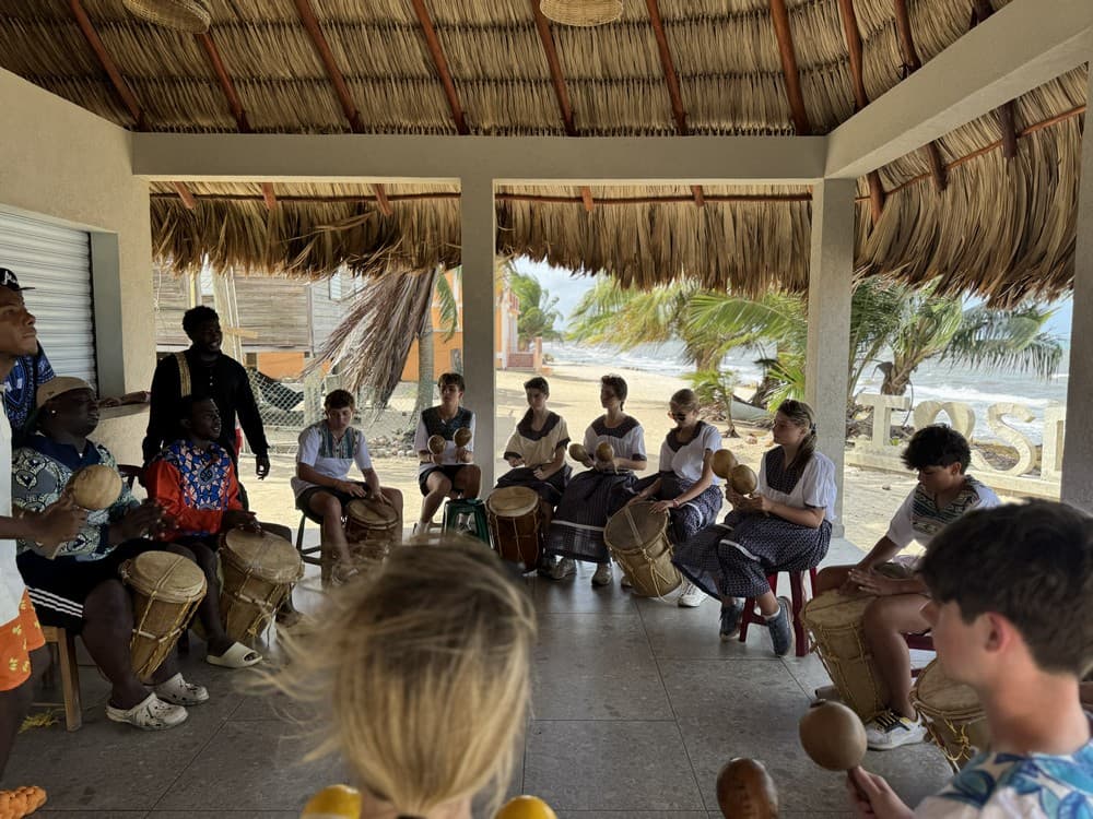 A group of people gathers under a thatched roof, playing drums and percussion instruments.