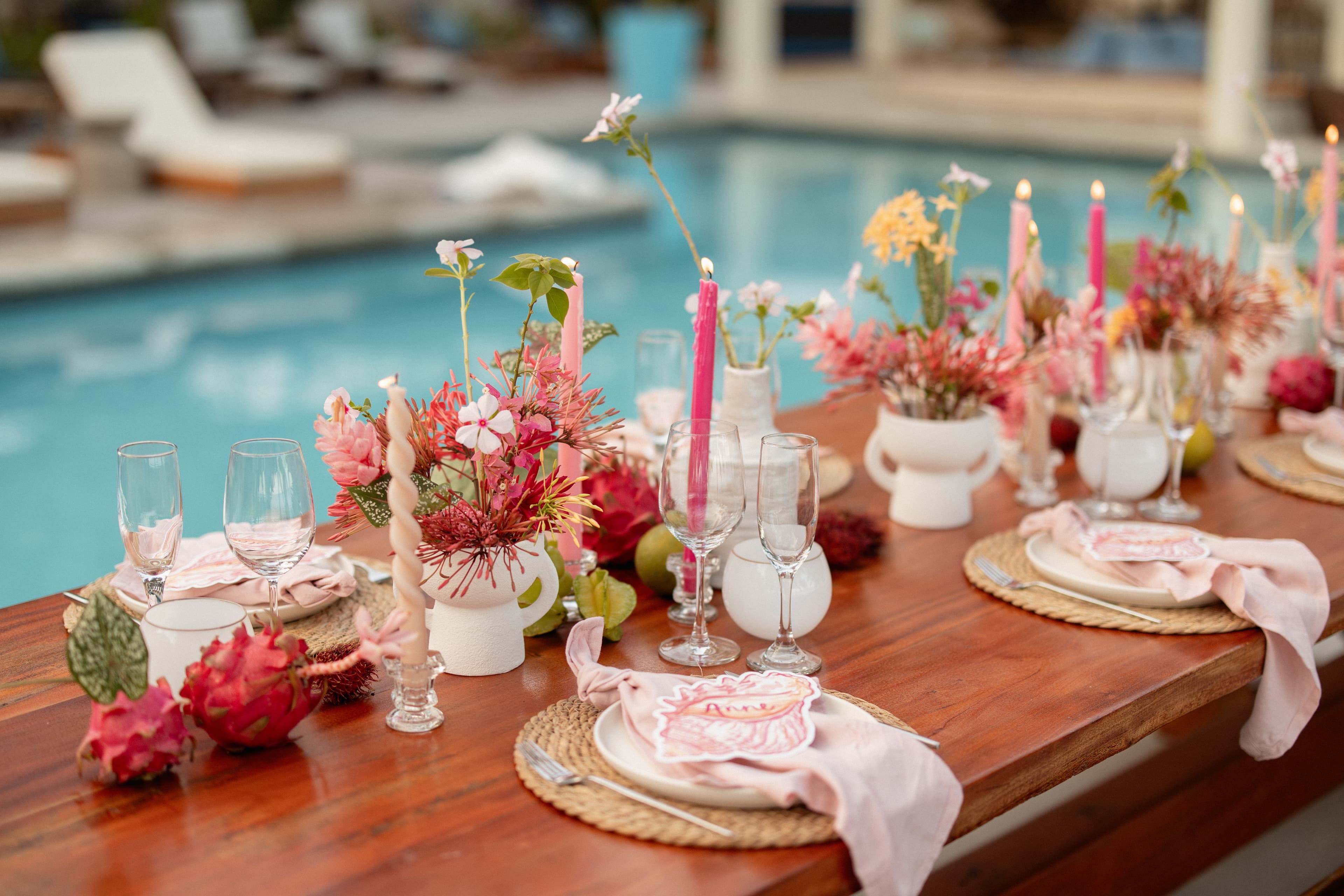 A beautifully set outdoor dining table adorned with pink flowers, candles, and glassware, beside a swimming pool.