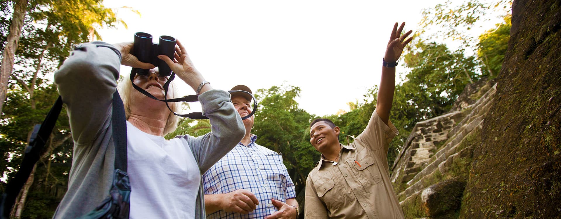 Three people engage in conversation at an archaeological site, with one using binoculars and another gesturing.