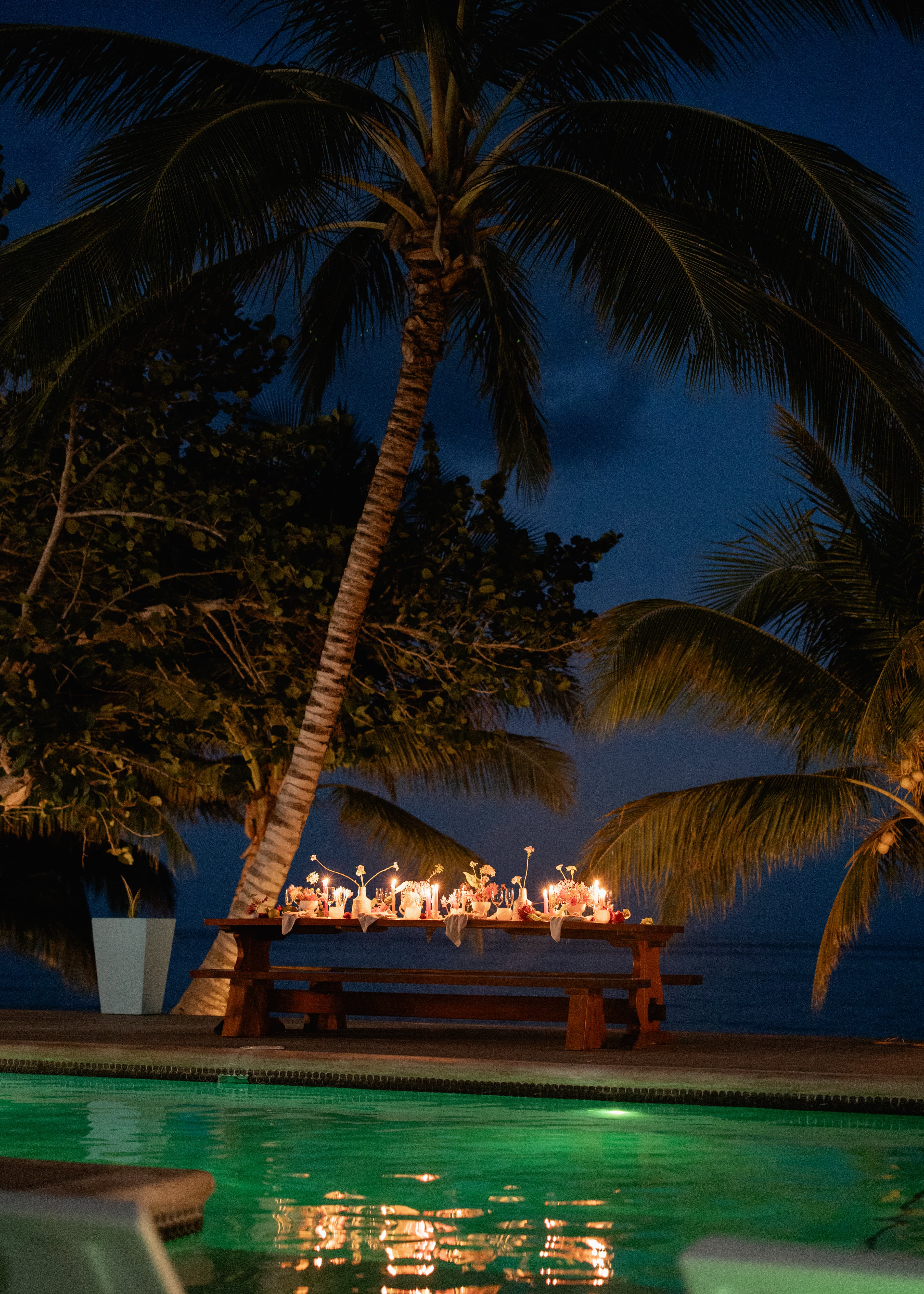 A beautifully set dining table with candles is illuminated by twilight near a tranquil pool and palm trees.