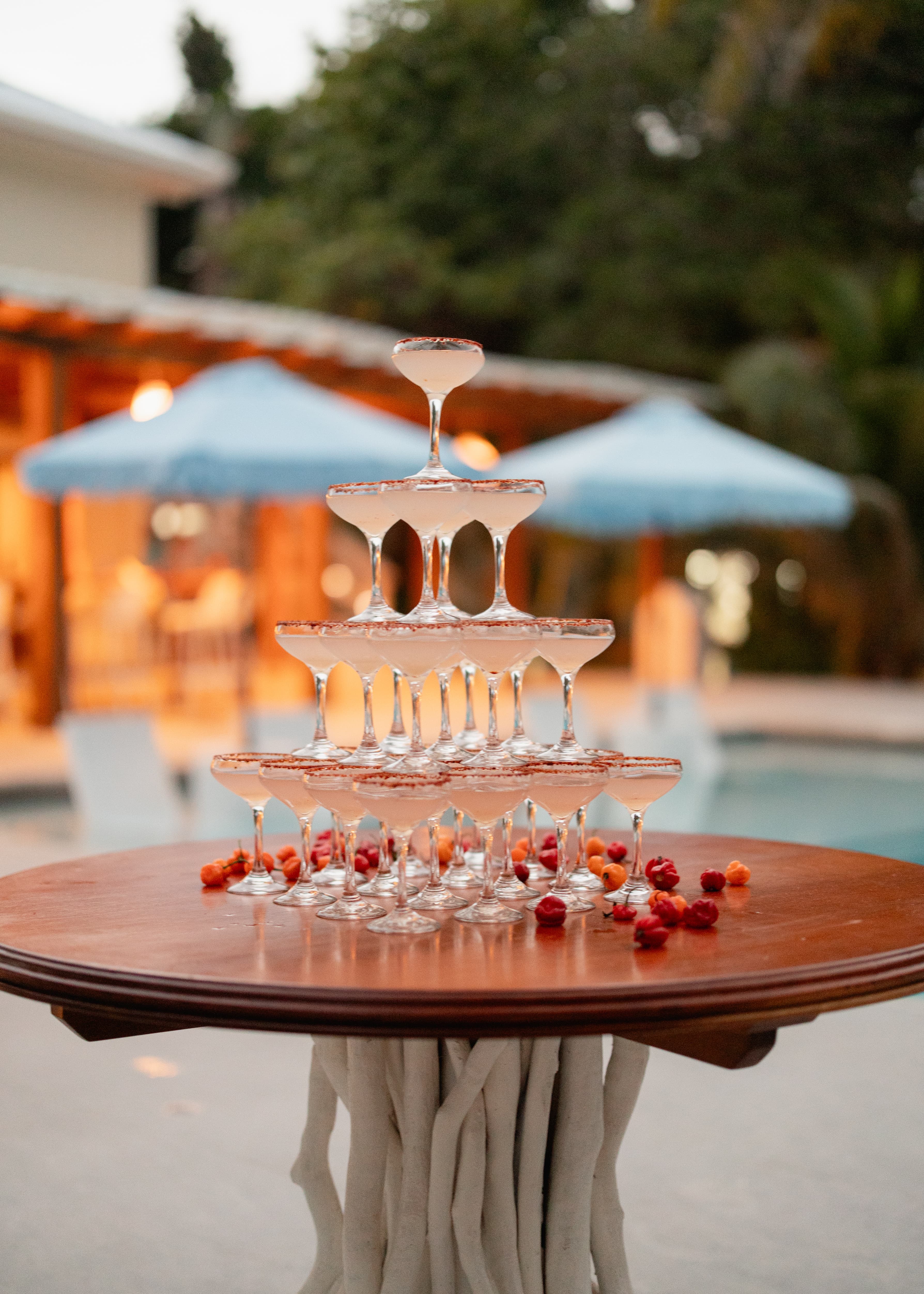 A tower of champagne glasses sits on a wooden table, surrounded by fruit, with a pool and umbrellas in the background.