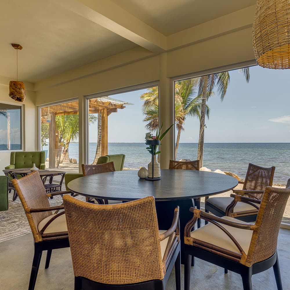 A dining area with a dark round table surrounded by chairs, featuring large windows overlooking a beach and palm trees.
