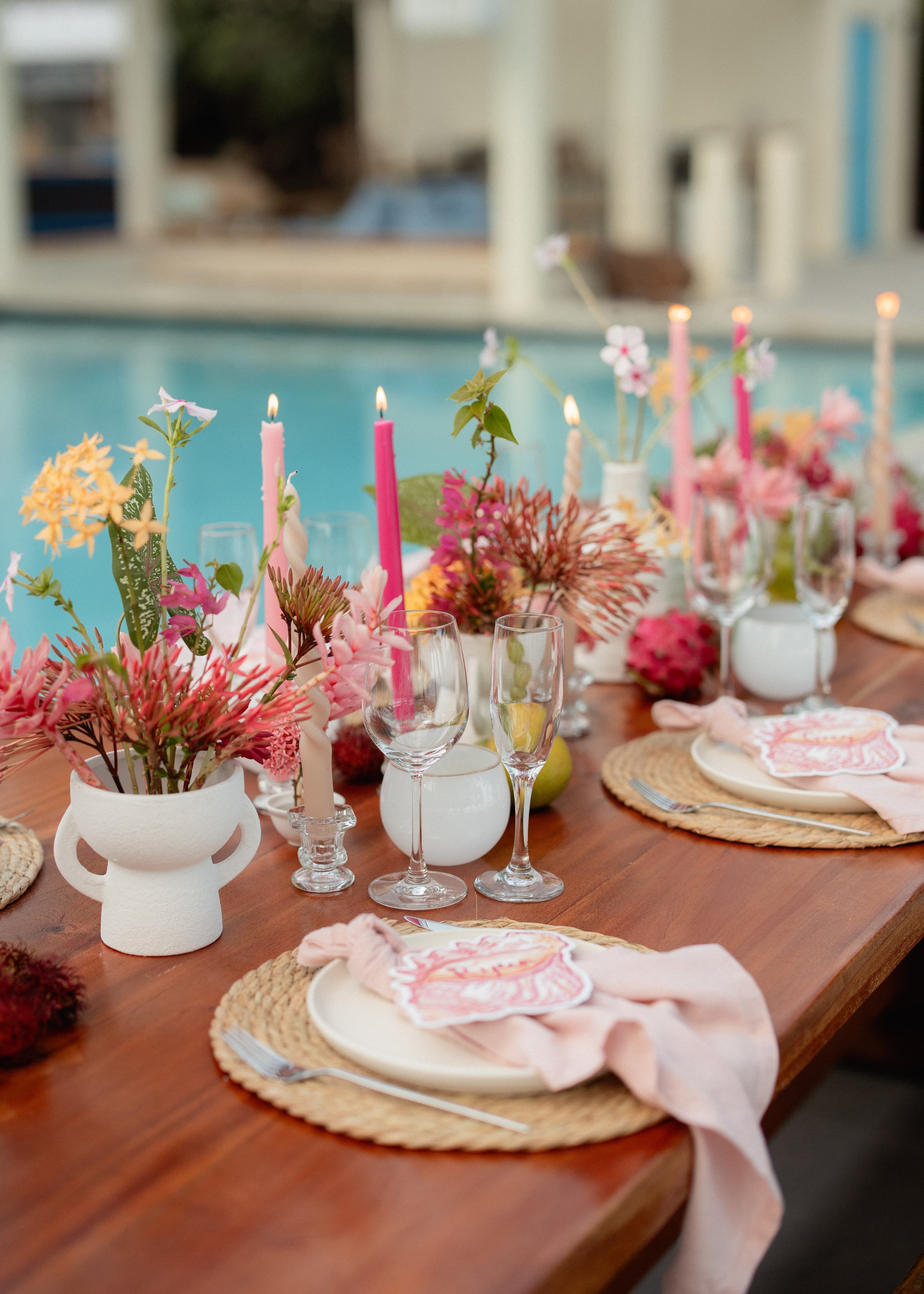 A beautifully arranged dining table with vibrant flowers, pink candles, and elegant dinnerware, set beside a swimming pool.