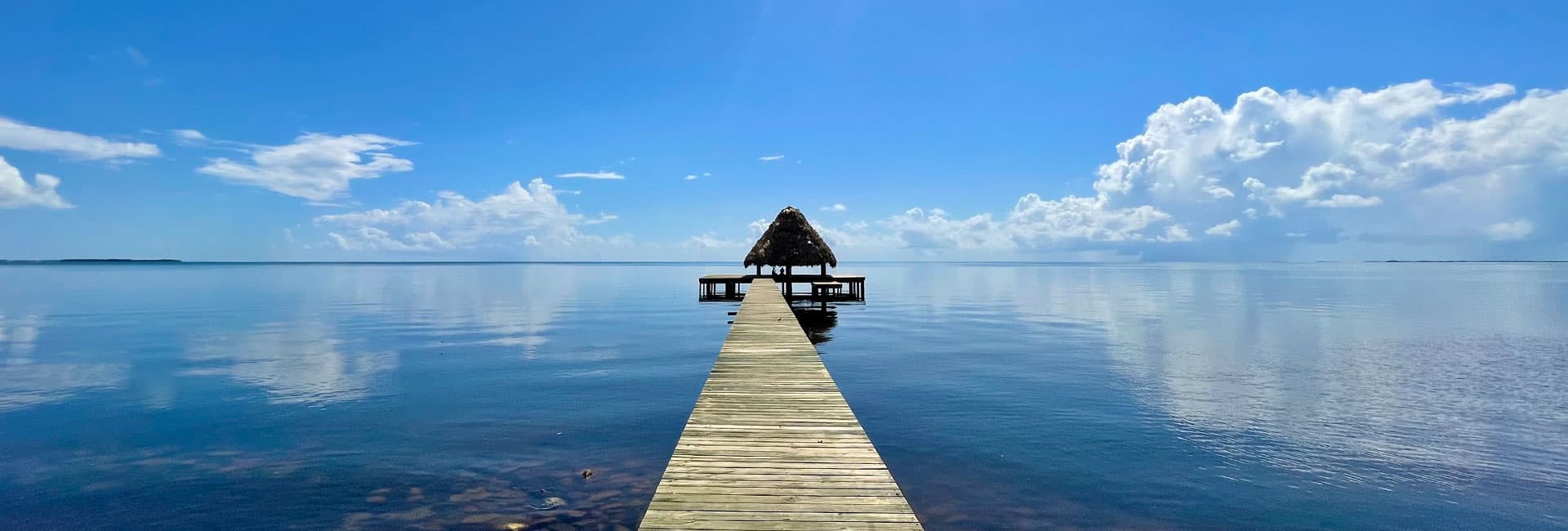 A wooden pier extends into calm blue waters under a clear sky with fluffy white clouds.