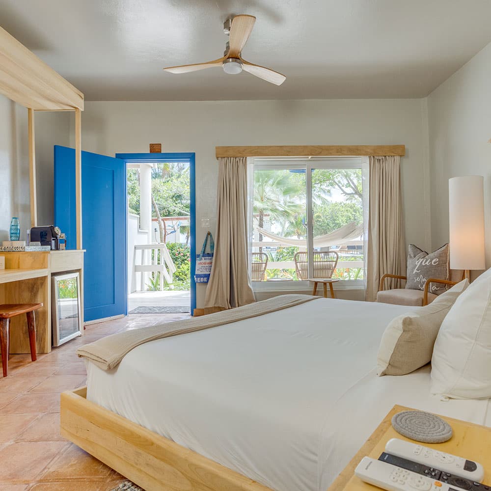 A bright, airy hotel room featuring a large bed, a writing desk, and a view of an outdoor patio through a blue door and window.