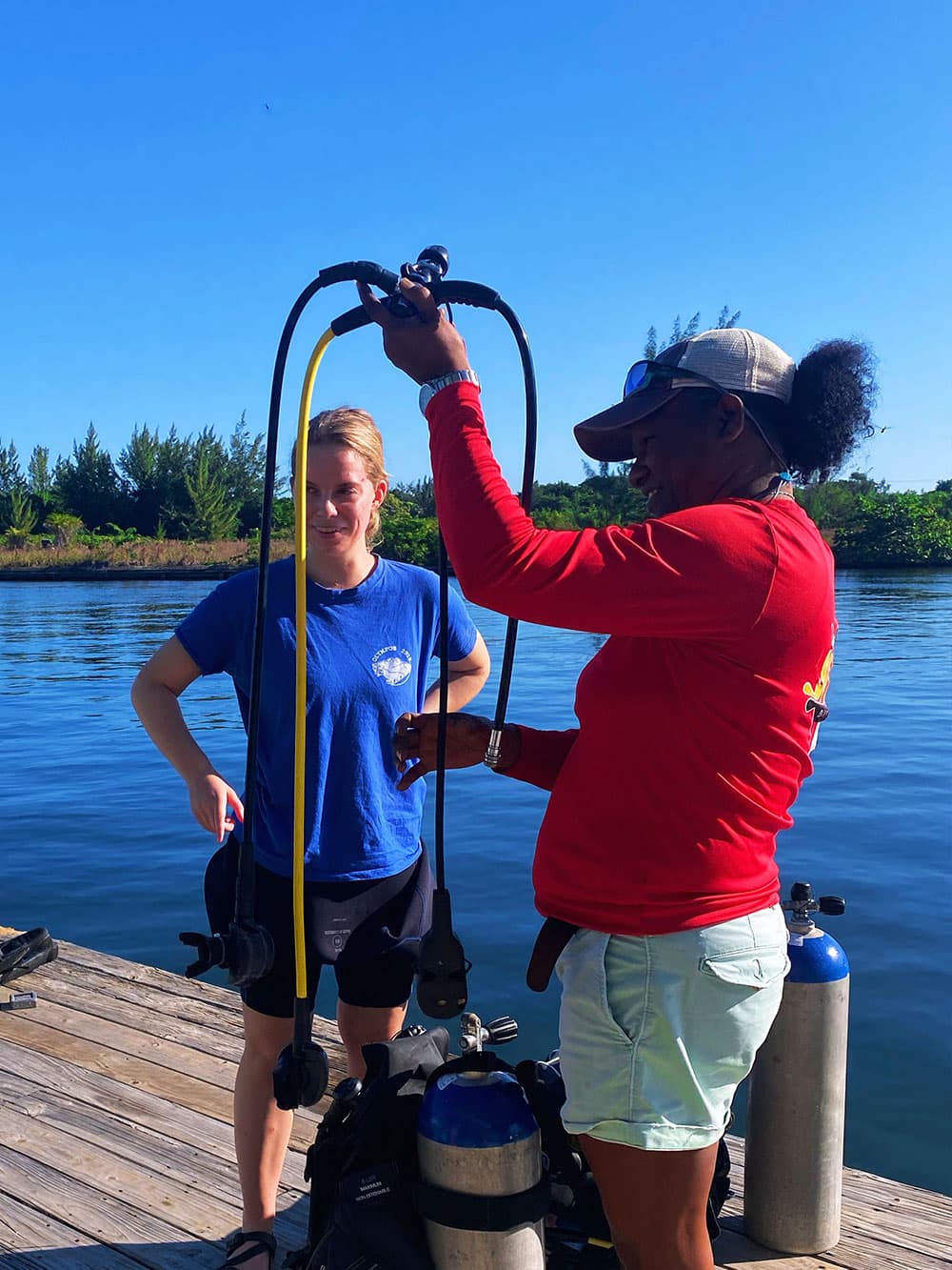 Two women prepare scuba gear by a dock on a sunny day.