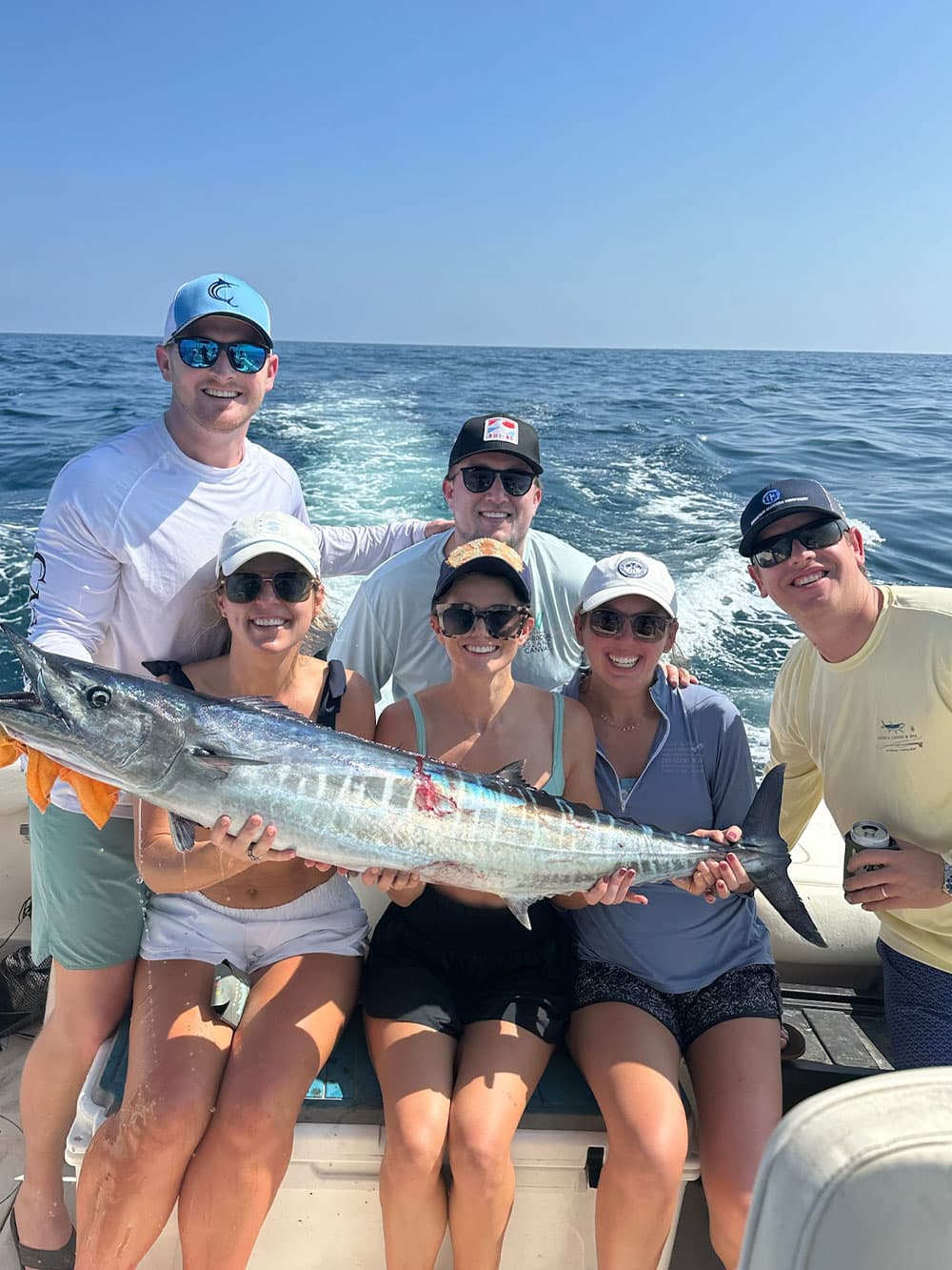 A group of six people on a boat proudly displaying a large fish against a clear blue sky.