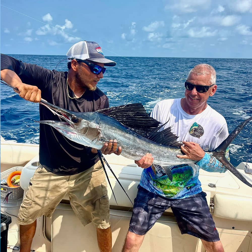 Two men on a boat hold up a large fish against a backdrop of ocean and sky.